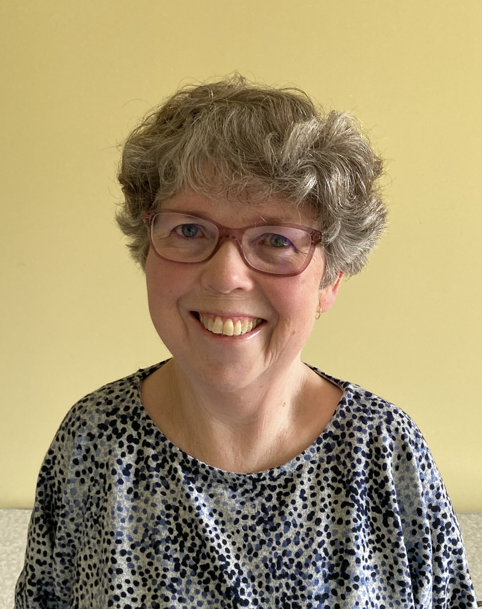 Trish Harris smiles broadly for a headshot in a dotty shirt and red classes.